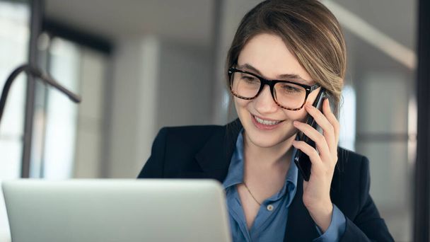 Młoda bizneswoman rozmawiająca przez telefon komórkowy i korzystająca z laptopa w biurze - Zdjęcie, obraz