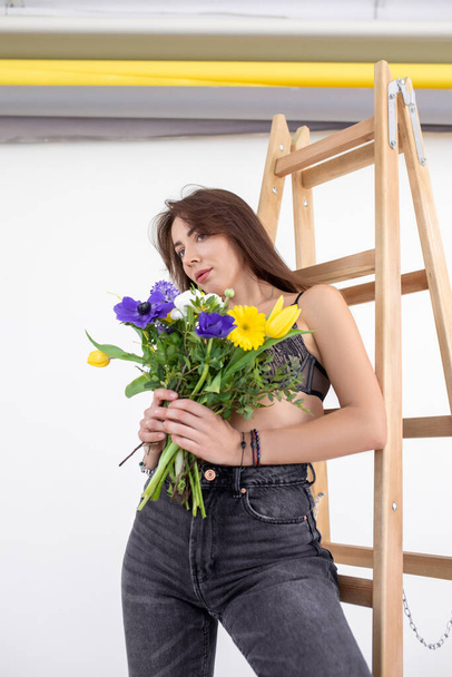 Portrait of graceful lady standing on ladder and covering breast with bouquet of bright flowers. Young woman with perfect makeup and stylish wear. Fashion photo, posing on white background in studio - Foto, Imagem