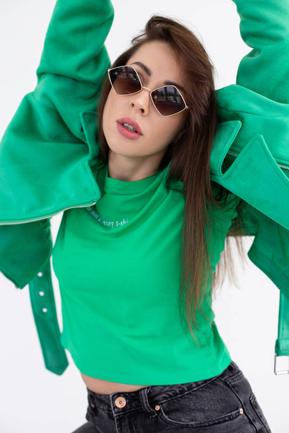 Portrait of young lady in black jeans, green jacket and t-shirt with sunglasses. Attractive woman with trendy accessory and stylish wear closeup. Fashion photo, posing on white background in studio - Фото, зображення