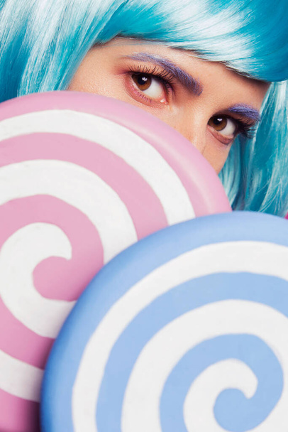 Beautiful sensual caucasian woman with exotic appearance in blue wig and colorful red lips, closing her with two big pink and blue lollypops on pink background - Valokuva, kuva