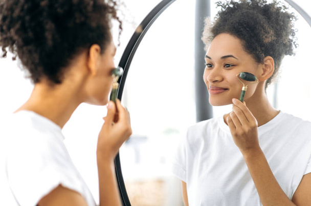 Chica encantadora afroamericana sosteniendo un rodillo de masaje de jade para la piel facial en su mano, hace medidas anti-envejecimiento, prevención de arrugas, de pie en casa frente al espejo, sonriendo - Foto, imagen