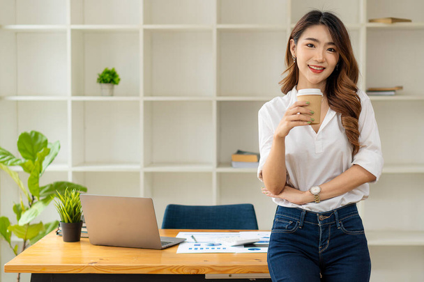 Young businesswoman, an Asian woman holding a cup of coffee standing in front of a desk in the office background with laptop and graph papers. - Photo, Image