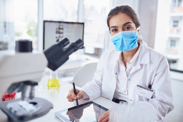 Nur ein weiterer Tag im Labor. Porträt einer selbstbewussten jungen Wissenschaftlerin, die eine chirurgische Maske trägt und an einem digitalen Tablet arbeitet, während sie in einem Labor in die Kamera blickt. - Foto, Bild