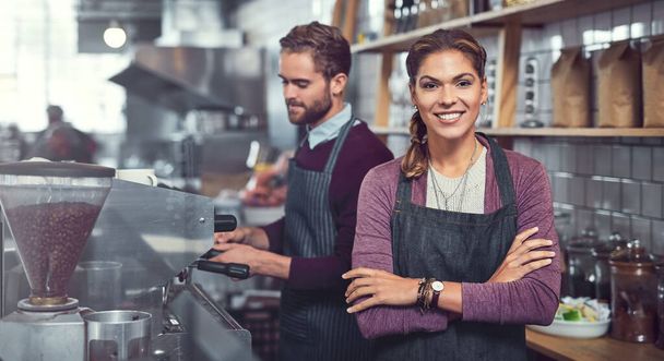 Existenzgründung - Leidenschaft sichtbar gemacht. Porträt einer selbstbewussten jungen Frau, die in einem Café arbeitet, während ihr Kollege im Hintergrund eine Kaffeemaschine bedient. - Foto, Bild