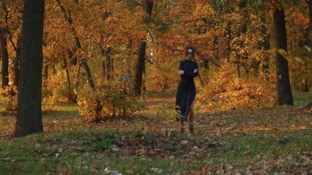 Una giovane donna con il trucco di Santa Muerte vestita con un vestito nero di morte cammina sullo sfondo delle foglie autunnali nella foresta durante il tramonto. Giorno dei morti o concetto di Halloween. - Filmati, video