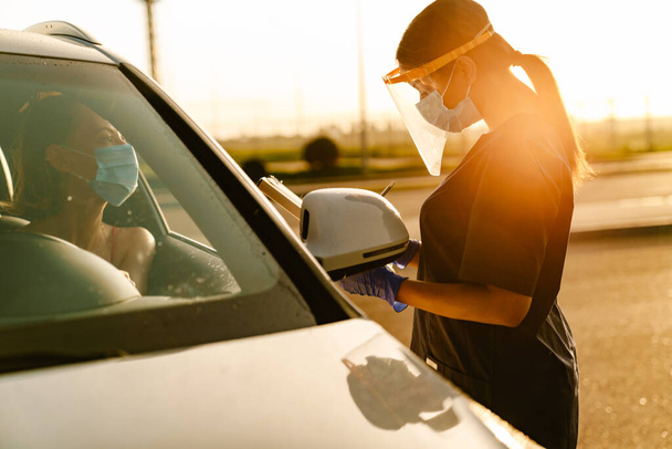 Black health worker in face mask writing down notes while talking car driver - 写真・画像