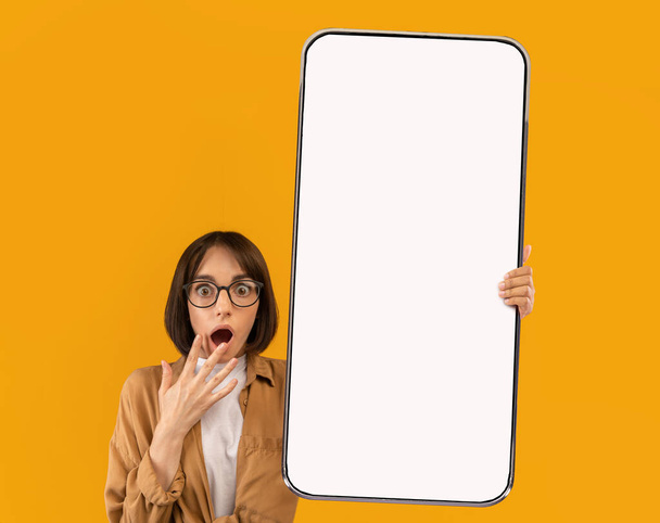 Mujer joven conmocionada en gafas que muestran un gran teléfono celular con pantalla en blanco, la publicidad de la nueva aplicación móvil, que ofrece maqueta - Foto, Imagen