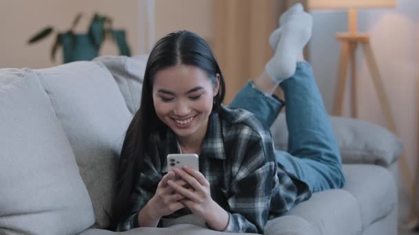 Азиатская корейская девушка японская китаянка лежала на диване, улыбаясь, глядя на мобильный телефон, смотрящий видео онлайн чаты сайт знакомств приложение SMS смартфон с бойфрендом заказ доставки - Кадры, видео