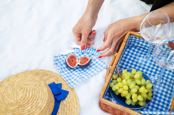 Romantic picnic in the park on the grass, delicious food: basket, wine, grapes, figs, cheese, blue checkered tablecloth, two glasses of wine. Girl cuts figs.Outdoor recreation concept - Photo, Image