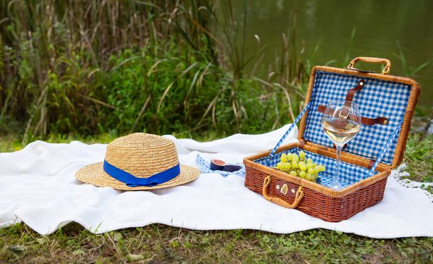 Romantische picknick in het park op het gras tegen de achtergrond van een prachtig meer, heerlijk eten: picknickmand, wijn, druiven, vijgen, kaas, blauw geruite tafelkleed, twee glazen wijn. - Foto, afbeelding