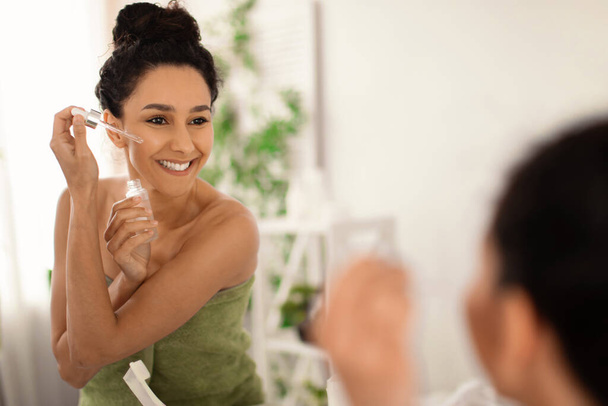 Jovem morena atraente aplicando soro hidratante em seu rosto perto do espelho em casa, espaço livre - Foto, Imagem