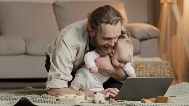 Svobodný otec vousatý muž táta s malou holčičkou dcera syn dítě dítě novorozenec sedí doma na podlaze při pohledu na notebook video hovor procházení on-line používá počítač app koupí dítě produkty on-line - Záběry, video
