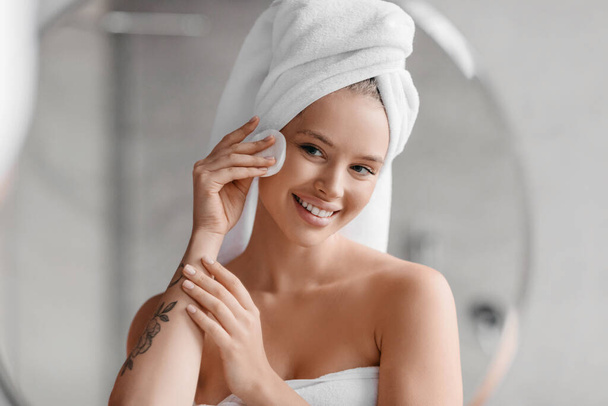 Soirée beauté routine. Belle femme nettoyant son visage avec du coton à la maison, portant une serviette sur la tête après le bain - Photo, image
