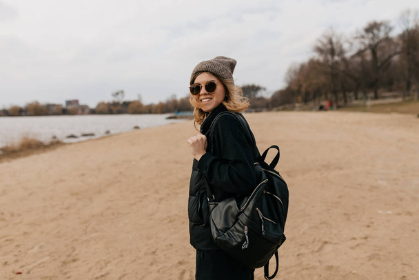 Excited lovely pretty girl with short hairstyle wearing cap with backpack in sunglasses is walking on the beach. Outside portrait of European woman enjoying walking. High quality photo - Foto, Imagem