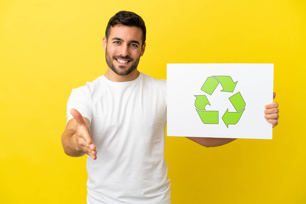 młody przystojny biały mężczyzna odizolowany na żółtym tle trzymając tabliczkę z recykling ikona co umowa - Zdjęcie, obraz