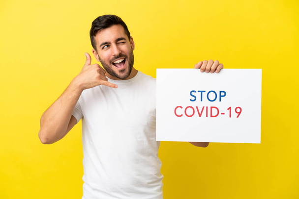 Young handsome caucasian man isolated on yellow background holding a placard with text Stop Covid 19 and doing phone gesture - 写真・画像