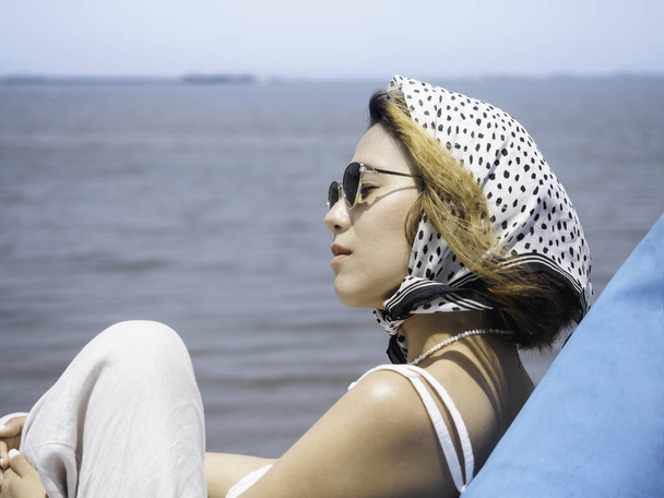 Magnifique portrait de femme asiatique avec les yeux fermés dans une chemise blanche décontractée portant des lunettes de soleil et une écharpe de cheveux assis sur un siège de sac de haricot bleu sur la plage avec vue sur la mer et fond bleu ciel en été. - Photo, image