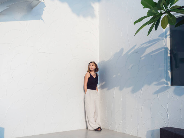 Asian woman look cool with short hair wearing black tank top and white trousers standing and keep hands in pockets on white building background, full range. Female summer fashion minimal style. - Photo, Image