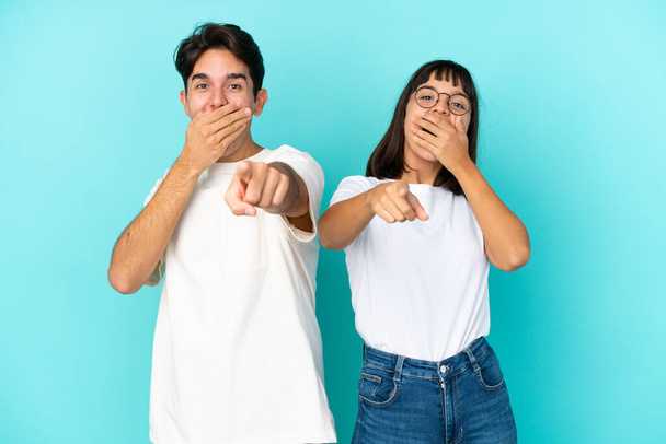 Nuori sekarotuinen pariskunta sinisellä taustalla osoittaa sormella jotakuta ja nauraa - Valokuva, kuva