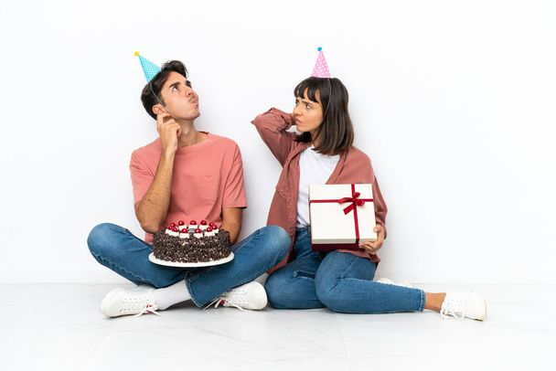 Fiatal vegyes faj pár ünnepli születésnapját ül a padlón elszigetelt fehér háttér gondolkodás egy ötlet, miközben vakarás fej - Fotó, kép