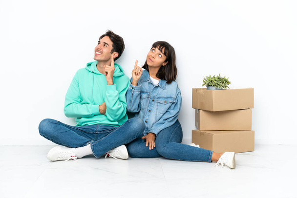 Молодая пара делает шаг, поднимая коробку с вещами, сидя на полу изолированы на белом фоне указывая на отличную идею и глядя вверх - Фото, изображение