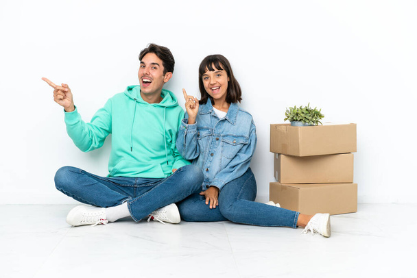 Молодая пара делает шаг, поднимая коробку с вещами, сидя на полу изолированы на белом фоне указывая пальцем в сторону и представляя продукт - Фото, изображение