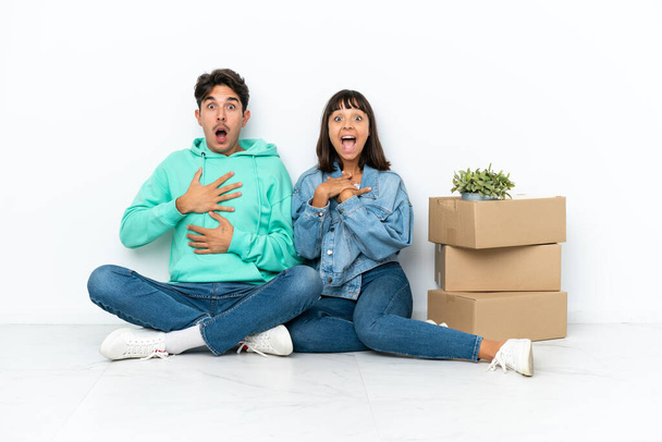 Молодая пара делает шаг, поднимая коробку с вещами, сидя на полу изолированы на белом фоне удивлен и шокирован, глядя прямо - Фото, изображение