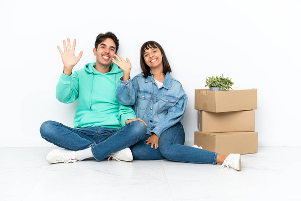 Молодая пара делает шаг, поднимая коробку с вещами, сидя на полу изолированы на белом фоне салютуя с руки со счастливым выражением - Фото, изображение