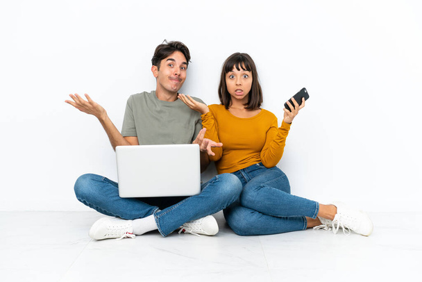 Молода пара з ноутбуком і мобільним пристроєм, що сидить на підлозі, має сумніви під час підняття рук і плечей
 - Фото, зображення