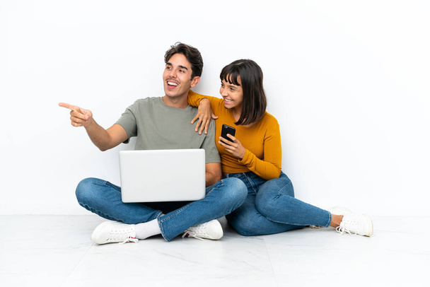 Молодая пара с ноутбуком и мобильным телефоном сидит на полу, представляя идею, глядя улыбаясь в сторону - Фото, изображение