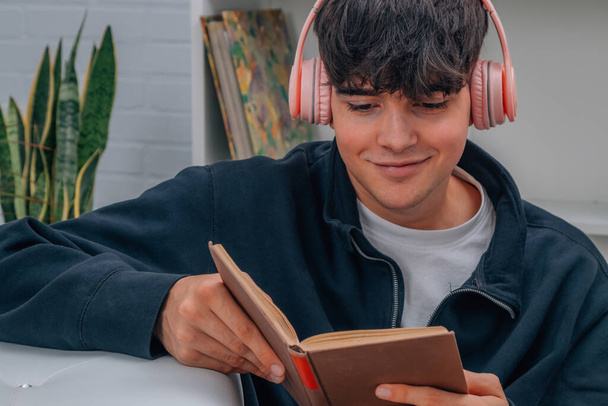 νεαρός άνδρας στο σπίτι διαβάζοντας βιβλίο και ακούγοντας μουσική με ακουστικά - Φωτογραφία, εικόνα