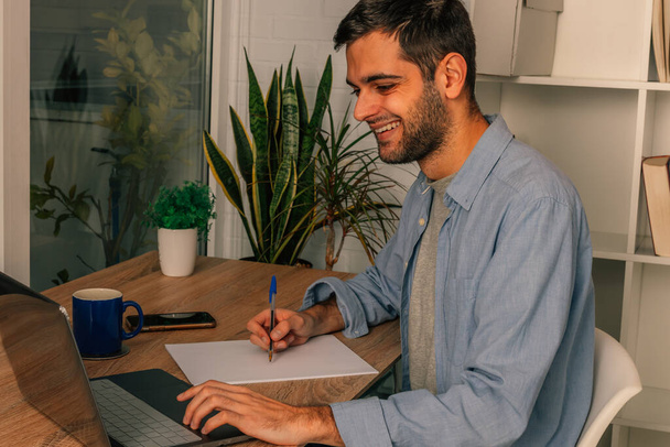 νεαρός άνδρας στο σπίτι με το γράψιμο του υπολογιστή ή κρατώντας σημειώσεις - Φωτογραφία, εικόνα