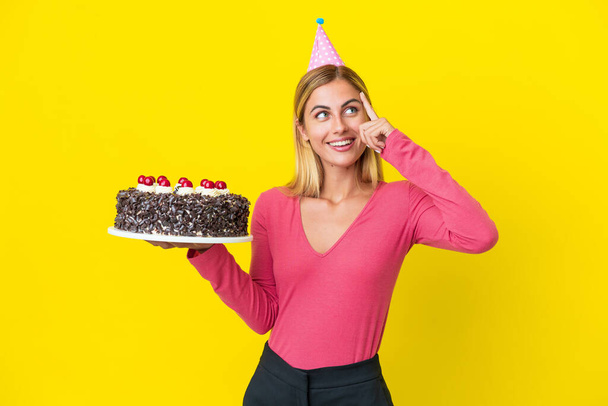 Блондинка Уругвая держит торт на день рождения изолирован на желтом фоне, сомневаясь и думая: - Фото, изображение
