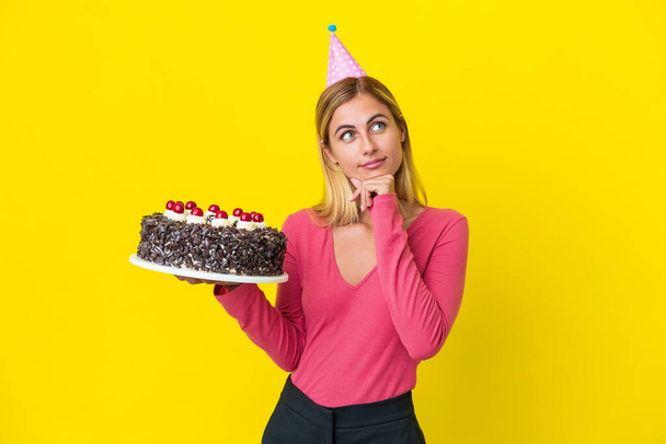 Chica rubia uruguaya sosteniendo pastel de cumpleaños aislado sobre fondo amarillo y mirando hacia arriba - Foto, imagen