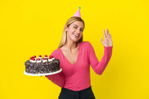 Blonde Uruguayenne fille tenant gâteau d'anniversaire isolé sur fond jaune saluant avec la main avec expression heureuse - Photo, image