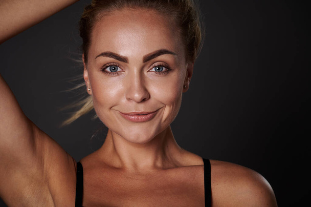Close-up portrait of the face of a beautiful woman with fresh clean glowing tanned skin, smiling looking at camera isolated over dark background. Body positivity, self-acceptance, confidence concept - Photo, Image