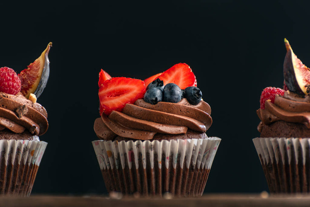Muffins on black background. Delicious chocolate cupcakes with cream decorated with strawberries and blueberries. Close up. - Fotoğraf, Görsel