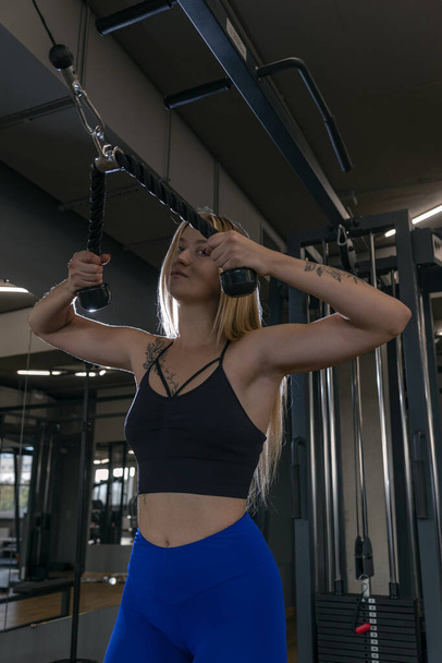 Πορτρέτο μιας όμορφης λεπτής γυναίκας στο γυμναστήριο. εκπαίδευση τρικεφάλων σε μηχάνημα γυμναστικής αντοχής. - Φωτογραφία, εικόνα