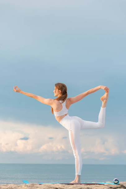 Młoda kobieta w białym stroju sportowym ćwiczy jogę rano na wybrzeżu. Uprawiam asanę Natarajasanę. Joga na plaży - Zdjęcie, obraz