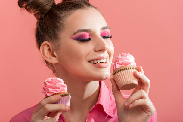 Girl with professional pink makeup enjoys delicious cupcakes. Portrait of woman with sweets on pink background - Zdjęcie, obraz