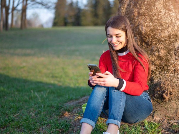 An attractive young brunette woman is sitting at the foot of a tree at the park. She gets a message.. Concept of carefree, communication use technology - Foto, Imagen