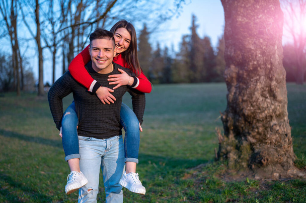 Couple caucasien dans le parc de printemps souriant, attrayant et heureux câlins fille beau gars sportif des épaules. Il se déplace hloding son piggyback, couple à l'extérieur. - Photo, image