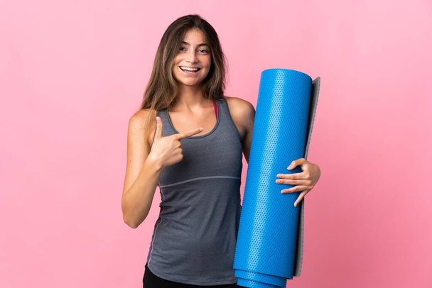 Молода спортивна жінка ходить на заняття з йоги, тримаючи килимок ізольований на рожевому фоні і вказуючи на нього
 - Фото, зображення
