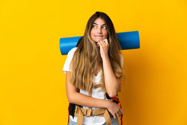 Young mountaineer woman with a big backpack isolated on yellow background having doubts and thinking - Photo, Image