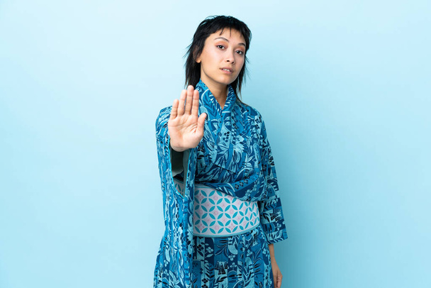 Mujer joven usando kimono sobre fondo azul aislado haciendo gesto de parada y decepcionado - Foto, imagen