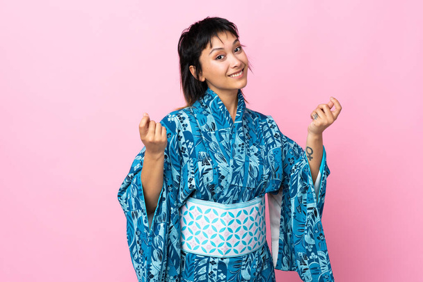 Молодая женщина в кимоно на изолированном синем фоне зарабатывает деньги жест, но разрушена - Фото, изображение