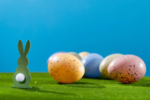 Easter holiday. Wooden rabbit decor. Colored spring eggs on green grass. Blue background. - Photo, image
