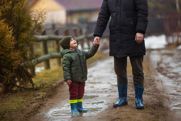 Baba ve oğul temiz havada, su birikintilerinde lastik botlarla yağmurdan sonra çamurda yürüyorlar. Küçük çocuk bir adamın elini tutuyor. - Fotoğraf, Görsel