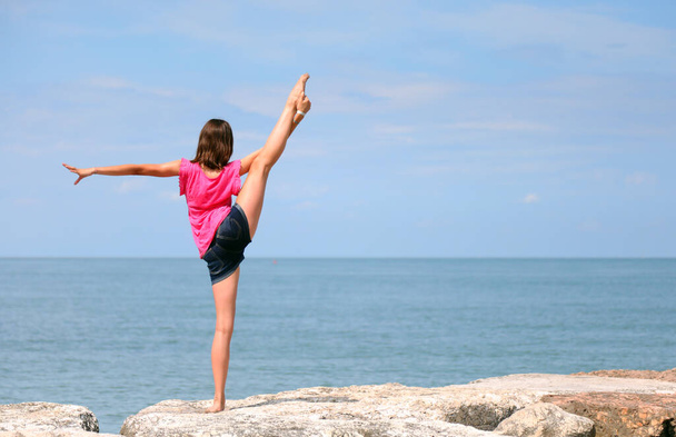 струнка молода дівчина з короткими штанами виконує ритмічну гімнастичну вправу, піднімаючи ногу над головою біля моря влітку
 - Фото, зображення