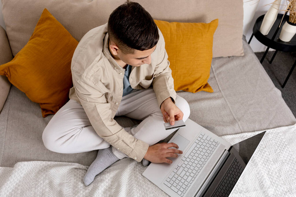 Junger Mann in legerer Kleidung surft auf seinem Laptop im Internet und hält eine Kreditkarte in der Hand, um etwas online zu kaufen. Einfaches Online-Shopping zu Hause, E-Commerce. Gemütlicher Innenraum. - Foto, Bild